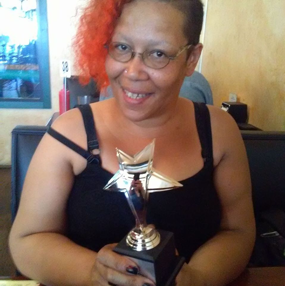Author Sumiko Saulson (an African American nonbinary person with orange hair) holding a star shaped Mixy Award in hir hands